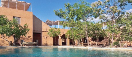 Sandfärgad fortbyggnad med träd och swimmingpool i förgrunden.