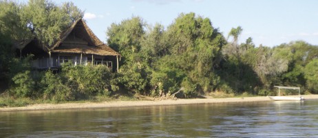 En tältbyggnad med spetsigt halmtak insprängd i grönskan vid flodstranden. I vattenbrynet ligger en båt med solskyddstak.